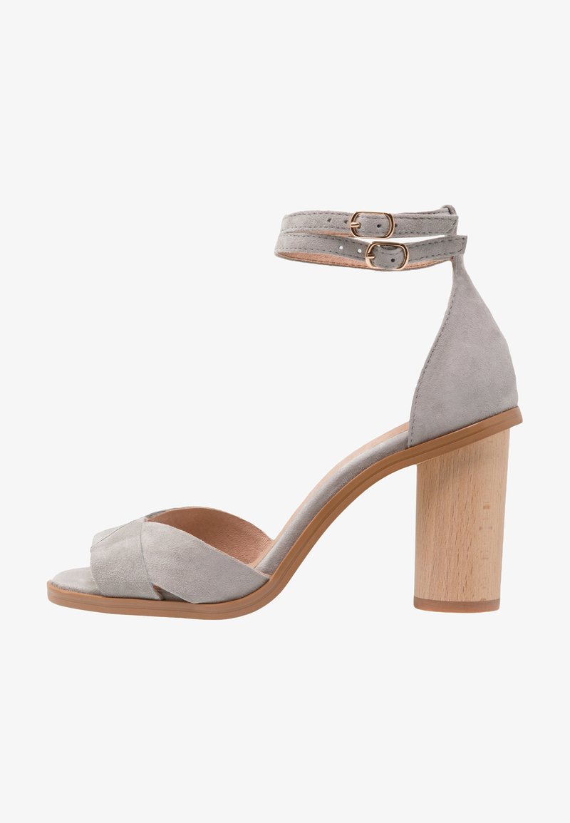 Sandalo con tacco alto in suede grigio con design a cinturino incrociato, chiusura con fibbia alla caviglia e tacco spesso in legno. Texture liscia su tutta la superficie.