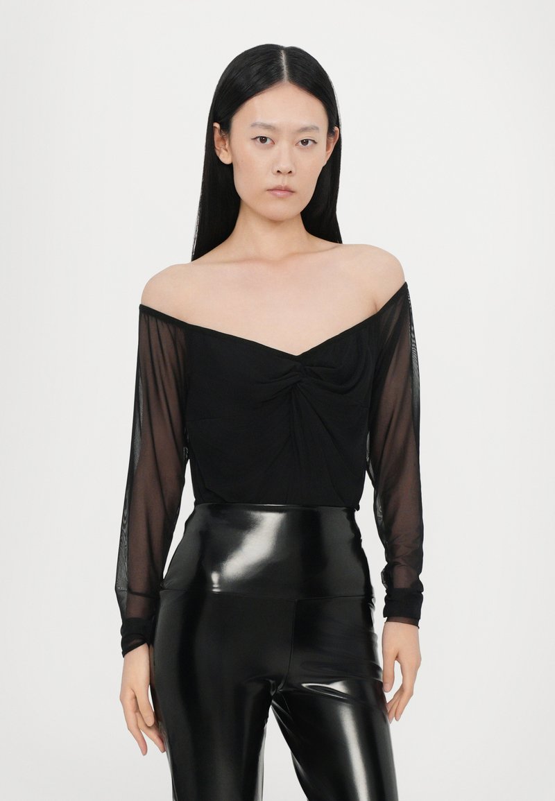 Top nero off-shoulder con maniche lunghe trasparenti e dettaglio frontale attorcigliato, abbinato a leggings neri lucidi a vita alta.