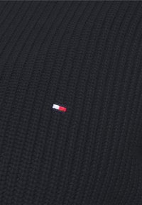 Mustaa ribbed-kudottua kangasta, jossa on pieni suorakulmainen punavalko-sininen brodeerattu logo keskellä.