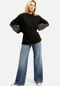 Czarny oversize sweter z szerokimi rękawami, zdobiony białymi spiralnymi wzorami. W połączeniu z jasnoniebieskimi spodniami o szerokich nogawkach i czarnymi sneakersami.