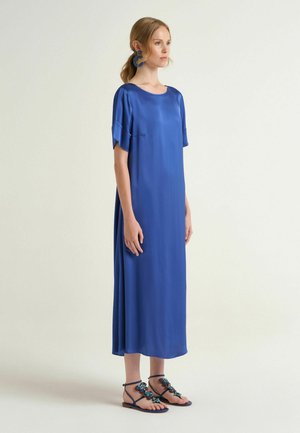 Donna in piedi con un vestito midi in satin blu con maniche corte e sandali bassi decorati con fiori di gioielli blu.