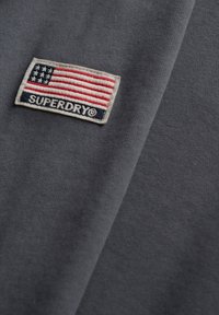 Grijze stof met een zachte textuur heeft een geborduurde patch met een rood, wit en blauwe vlag en de tekst "SUPERDRY."