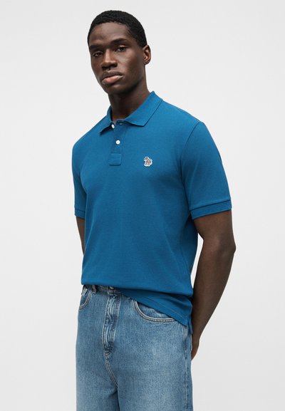 Μπλούζα πόλο από βαμβάκι σε τιρκουάζ απόχρωση, με κλασικό γιακά, κοντά μανίκια και μικρό κεντημένο λογότυπο στο αριστερό στήθος. Συνδυάζεται με ανοιχτόχρωμο τζιν.
