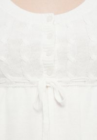 Λευκό πλεκτό πουλόβερ με υφασμένο σχέδιο καλωδίου, πέντε κουμπώματα και λεπτομέρεια δέσιμου στη μέση. Απαλή υφή με ριμπ μανσέτες.