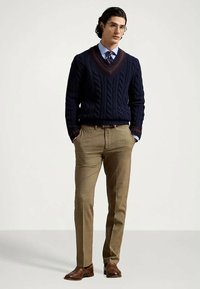 Άνδρας στέκεται με τα χέρια στις τσέπες, φορώντας σκούρο μπλε πλεκτό πουλόβερ, ανοιχτό μπλε πουκάμισο, γραβάτα, καφέ καρό παντελόνι και καφέ δερμάτινα παπούτσια.