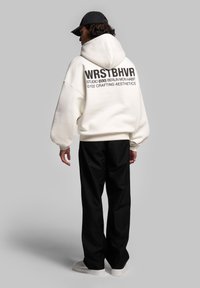 Felpa bianca oversize con cappuccio, con testo nero sul retro. Abbinata a pantaloni neri e sneaker bianche, su uno sfondo grigio.