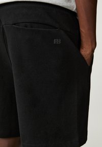 Schwarze Shorts aus strukturiertem Stoff, mit einer Seitentasche und einem kleinen schwarzen Logo am Bund. Einfaches und funktionales Design.
