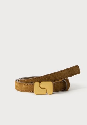 Une ceinture en daim marron enroulée, aux bords surpiqués et dotée d’une grande boucle dorée, sur un fond blanc uni.