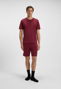 Burgundy t-shirt med kort ärm och minimalistisk logotyp, matchande shorts. Bärs med svarta ankelsockor. Slät textur, avslappnad passform.