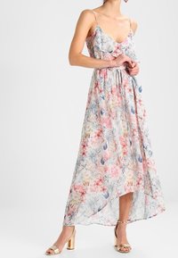 Abito maxi floreale con spalline sottili, tessuto leggero in colori pastello, presenta volant e una silhouette fluida, abbinato a décolleté in oro con tacco quadrato.