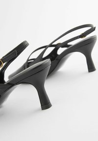 Musta nahkainen slingback-kengät, joissa on pyöreä muoto, ohuet hihnat ja pieni kultainen yksityiskohta kantapäässä. Rakenteeltaan teksturoitu pinta ja matala lohkokorko.
