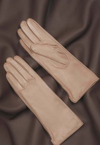 Beige leather gloves with a smooth texture, featuring a fitted design and raised stitching detail along the wrist area, laid on brown fabric.