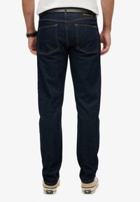 Los jeans de mezclilla azul oscuro presentan un corte recto, costuras naranjas en contraste, dos bolsillos traseros y una etiqueta de cuero con la marca en la cinturilla.