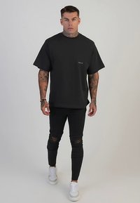 Schwarzes T-Shirt aus Baumwolle mit kurzen Ärmeln, einem Rundhalsausschnitt und einem kleinen weißen Logo. Dazu schwarze, zerrissene Skinny-Jeans und weiße Turnschuhe.