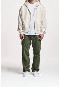 Felpa crema con zip, t-shirt bianca sotto, pantaloni cargo verde oliva con tasche laterali e sneaker bianchi. Tessuto liscio con vestibilità rilassata.