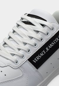 Λευκό δερμάτινο αθλητικό παπούτσι με λευκά κορδόνια και μαύρη ρίγα στο πλάι με την επιγραφή "Versace Jeans Couture".