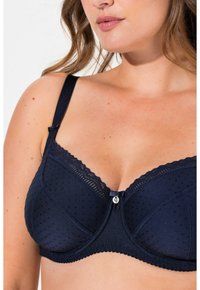 Tummansinisellä rintaliivillä on pilkkukuosi, pitsireunus ylhäällä, säädettävät olkahihnat ja keskellä koristeellinen nappi.