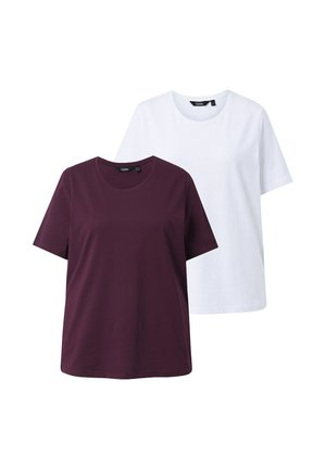 Frau mit schulterlangem, welligem blondem Haar, die ein einfaches, dunkelviolettes Kurzarmshirt und blaue Jeans trägt und vor einem weißen Hintergrund steht.