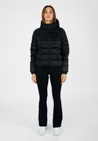Giacca a vento nera con cappuccio, caratterizzata da un design trapuntato, chiusura con zip e polsini elasticizzati. Indossata con pantaloni neri e sneakers bianche.
