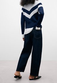 Pull en maille bleu marine avec motif chevron blanc ; associé à un jean en denim foncé ample et des chaussures noires à enfiler. Design minimaliste, sans logos visibles.