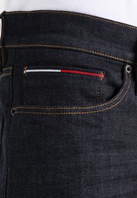 Mörka denimjeans med en bakficka som har röd, vit och blå sömnad, kontrasterande orange tråd och en metallnitar.