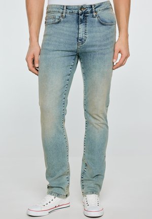 Mężczyzna w jasnoniebieskich dopasowanych dżinsach i białych trampkach, stojący na gładkim tle, z rękami swobodnie opuszczonymi wzdłuż ciała.