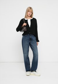 Schwarze knöpfbare Strickjacke, weißes T-Shirt, lockere blaue Jeans und helle Sneakers. Kleine schwarze Handtasche. Einfarbiger weißer Hintergrund.