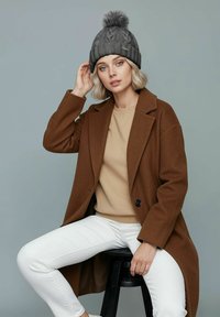 Brown wool coat, beige sweater, white pants, and a gray knitted beanie with a pom-pom. Model sitting on a black stool against a gray backdrop.