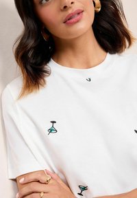 T-shirt en coton blanc avec des verres à cocktail brodés en noir et bleu. Encolure ronde avec une coupe décontractée et des manches courtes.