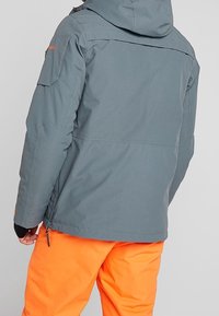 Veste imperméable grise avec une capuche, dotée d'une poche latérale, de poignets ajustables et d'une coupe droite, assortie à un pantalon orange vif.