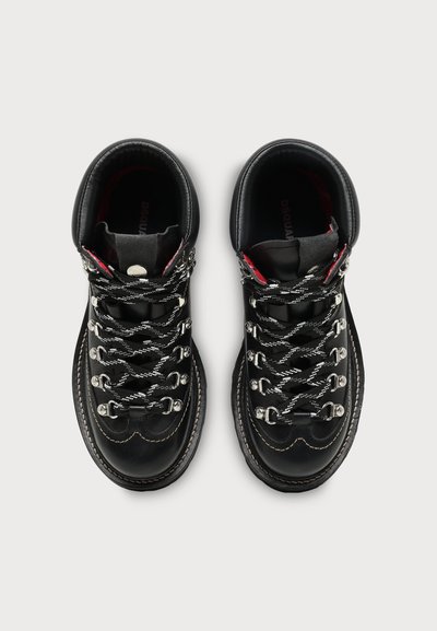 Paire de bottes de randonnée en cuir noir avec des lacets noirs et blancs, des œillets métalliques et des coutures visibles sur un fond clair uni.