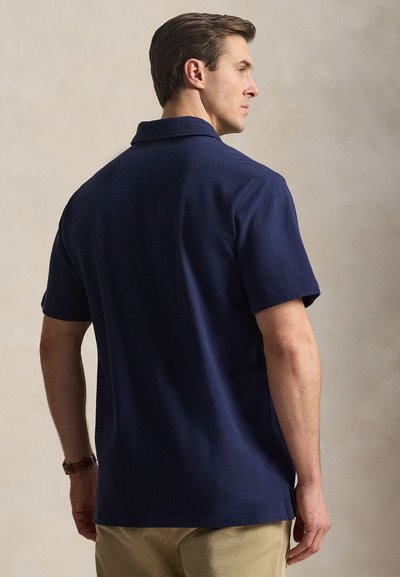 Άνδρας που φορά μπλε σκούρο πόλο μπλουζάκι με κοντά μανίκια και μπεζ παντελόνι, γυρισμένος πλευρικά σε απλό μπεζ φόντο.