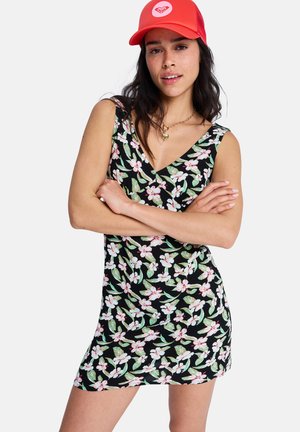 Czarna sukienka w kwiaty z różowymi i zielonymi wzorami, z dekoltem w serek i dopasowanym fasonem. Modelka ma na sobie jasnoczerwoną czapkę.