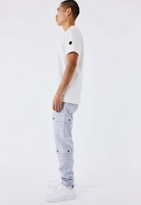 Giovane in t-shirt bianco, pantaloni cargo grigio chiaro con bottoni neri e sneakers bianche, in piedi di profilo contro uno sfondo bianco semplice.