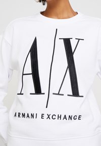 Felpa bianca con un logo ricamato nero in evidenza che riporta la scritta "A/X" e "ARMANI EXCHANGE" sul petto. Tessuto morbido con scollatura rotonda.
