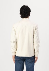 Lys beige jakke i et glat, let shiny stof med opretstående krave og lange ærmer, parret med blå jeans. Bagansigt vist.