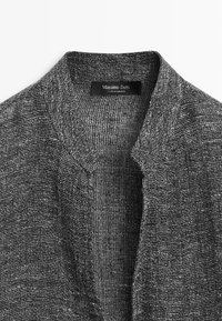 Col de cardigan en maille texturée grise avec une étiquette noire indiquant Massimo Dutti made in Morocco sur fond blanc.