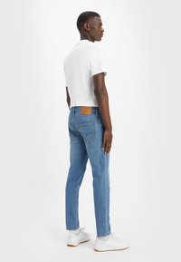 Modri jeans s slim fit krojem, z zadnjimi žepi, usnjenim logotipom na pasu, v kombinaciji z belo majico s kratkimi rokavi in belimi supergami.