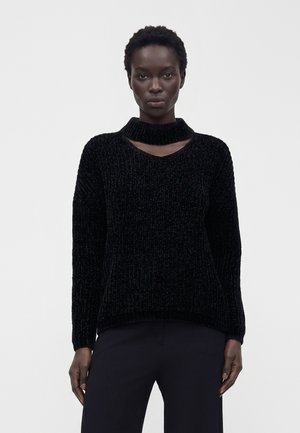 Femme à la peau foncée, debout face à l’avant, portant un pull noir texturé à manches longues avec une découpe col ras-du-cou et un pantalon noir.
