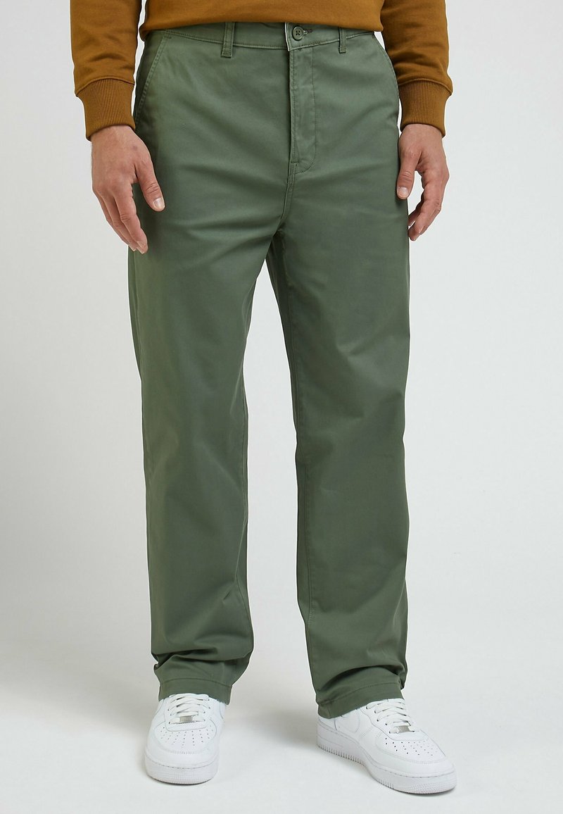 Personne portant un pantalon vert olive, des baskets blanches et une chemise marron à manches longues, debout devant un fond uni.