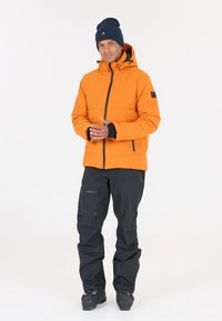 Veste isolante orange avec fermeture éclair sur le devant, accents noirs et capuche ajustable, associée à un pantalon noir et des chaussures de ski. Design simple et fonctionnel.