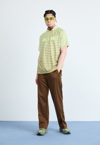 Polo shirt rayé vert clair avec le texte "AIR", associé à un pantalon marron et des baskets à motifs verts. Des lunettes de soleil claires complètent le look.