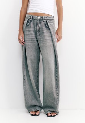 Osoba oblečená v sivých džínsoch s vysokým pásom a širokými nohavicami a čiernych žabkách, v kombinácii s bielym krátkym tielkom.