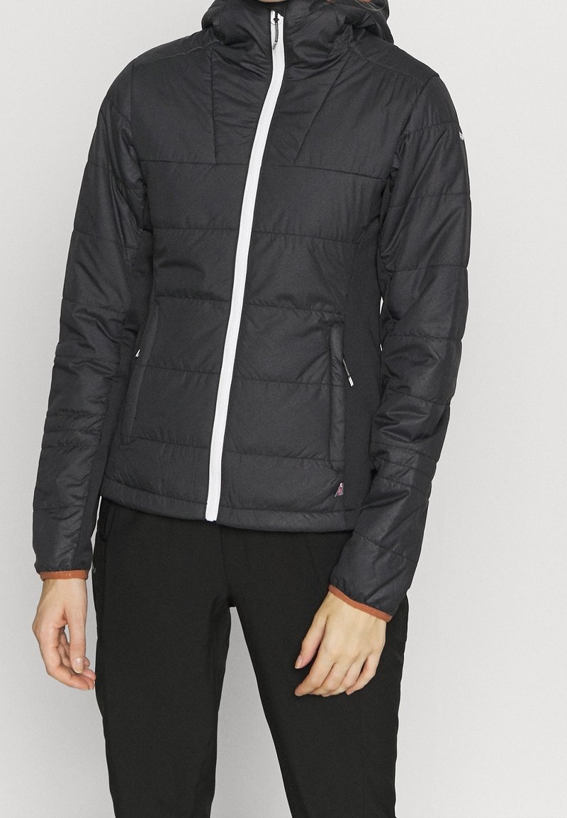 Personne portant une veste matelassée noire avec une fermeture éclair blanche, des poches latérales et une capuche, associée à un pantalon noir.