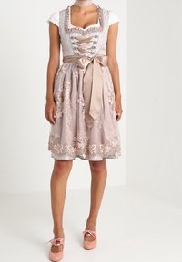 Robe dirndl avec un corsage blanc, un ruban décoratif rose et une jupe brodée longueur genou avec des motifs floraux. Accompagnée de chaussures roses.