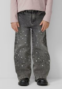 Dziecko w czarnych dżinsach z rozsianymi srebrnymi cekinami i czarnych błyszczących butach, stojące na jednolitym szarym tle.