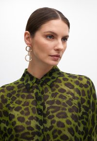 Zelená leopardia potlač priesvitná košeľa s golierom, zapínaním na gombíky a čiernymi škvrnami; skombinovaná s veľkými zlatými kruhovými náušnicami.