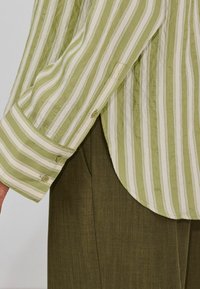 Camisa de manga larga a rayas verdes y blancas con bajo redondeado, puños abotonados y tejido texturizado. Combinada con pantalones verde oliva.