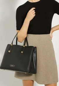 Black leather tote bag with gold accents, rectangular shape, textured surface, and sturdy handles. Displayed next to a beige skirt and black top.