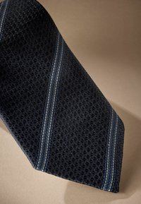 Cravatta blu navy con un motivo testurizzato e accenti a righe diagonali blu, caratterizzata da una finitura liscia e mantenendo una forma triangolare elegante.